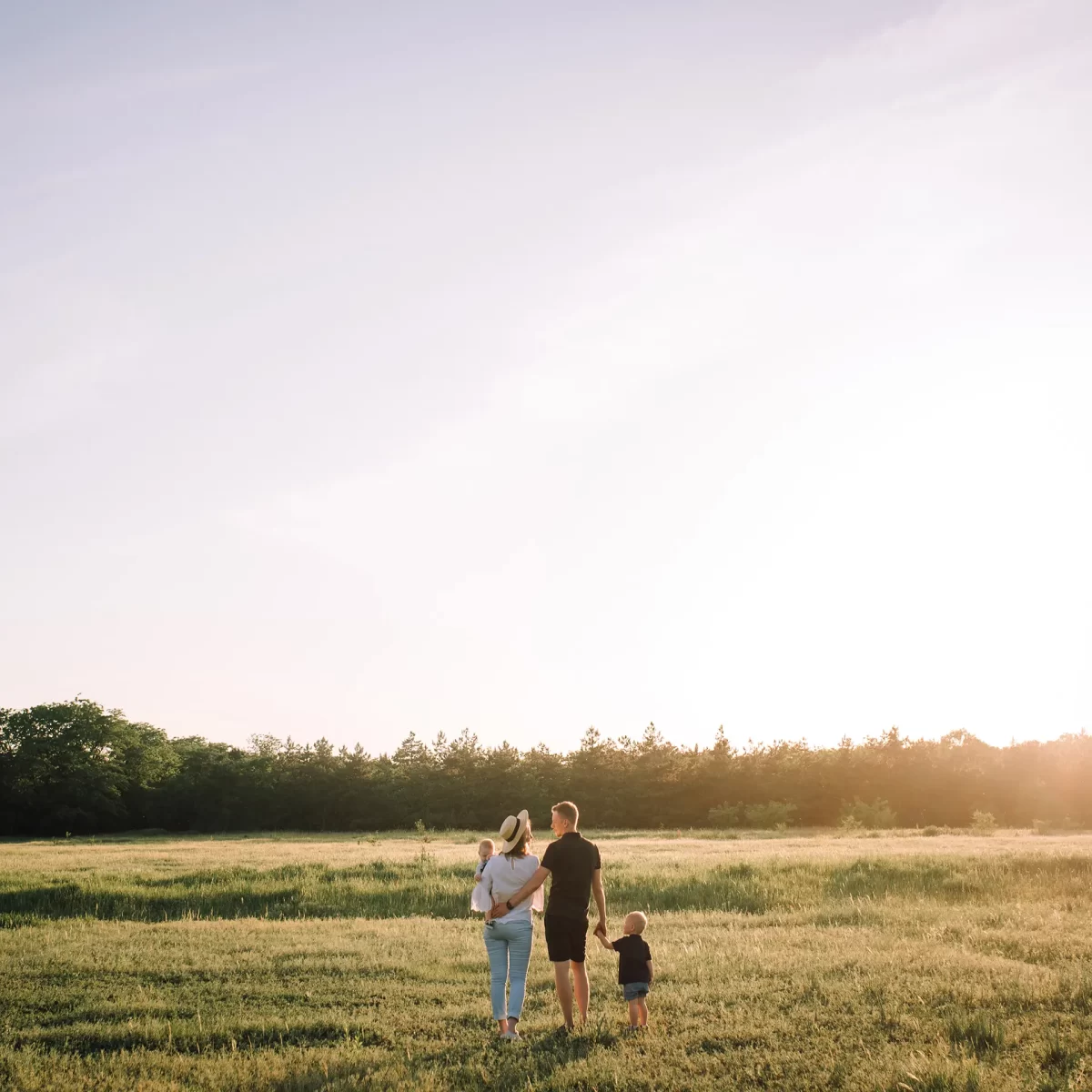 Family of four stands together in a grassy field at sunset, facing the horizon with trees in the background and a warm glow from the setting sun.